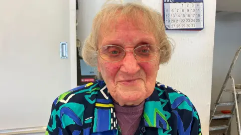 A close-up of an elderly woman with short blonde curly hair and glasses. She is wearing a patterned green, black, blue and yellow shirt over a purple long-sleeved top. Behind her is a white cupboard and wallpaper and a wall calendar.