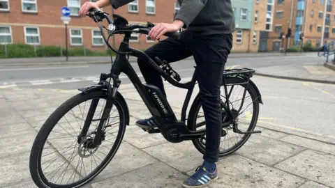 Simon Thake/BBC A man sitting on a black stationary e-bike