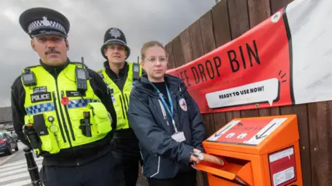 North Yorkshire Police Two police officers and a council officer stand next to an orange metal knife bin, with "knife drop bin" written on a large banner behind it. The council community safety officer is placing an object shaped like a blade but wrapped in cardboard into the bin.
