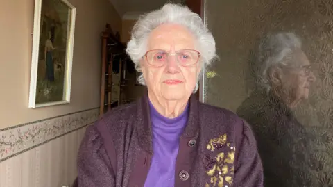 91-year-old Annie James stands in her doorway. She has silver short hair and wears large glasses with a reddish-brown rim. She wears a lilac purple turtleneck, with a dark purple cardigan over the top. 