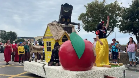 A Snow White themed float which has a huge red apple with a green leaf at the front of it. To its side stands a girl dressed as Snowe White, wearing a yellow skirt, a black top with red and black stripes and a red bow around her dark hair. Behind the apple is a girl kneeling, as if hiding, who is dressed as a witch and wears a black dress, black tights and a black cape with the hood over her eyes. Behind her is a yellow gabled house and a large fawn. Behind the float are men, women and children walking who are wearing tunics, hats and long red dresses respectively. There are spectators taking photos.