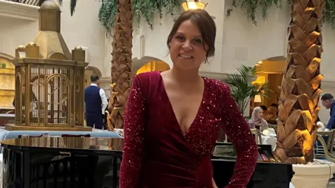 A woman with dark brown hair pinned up. She is wearing a long burgundy dress and behind her is a grand piano, palm tress and table and chairs. 