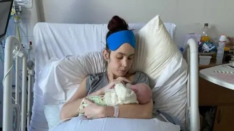 Natalie is sitting upright in a hospital bed, cradling a newborn baby wrapped in a patterned blanket and cream-coloured knitted hat. The hospital room contains medical equipment and bedside supplies, with pillows propped behind the patient and bedding covering most of the bed. A blue cooling eye mask rests on the person's forehead, while the baby is held close against their chest.