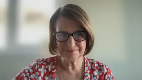 Patient Safety Commissioner Karen Titchener sits indoors facing the camera, wearing dark-framed glasses and a red patterned top, with a softly blurred background.
