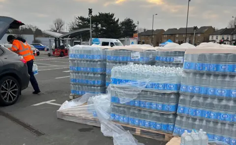 Man in high vis jacket stands at rear of car parked next to pallets with hundreds of bottles of water in plastic bottles wrapped in plastic sheets