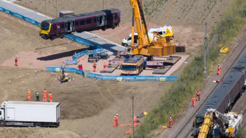 BAV MEDIA/GEOFF ROBINSON PHOTOGRAPHY A yellow crane is lifting the purple train with a winch. It is seen hanging over a large brown field, with lots of workers wearing orange clothing below. The other train is still on the tracks on the right.