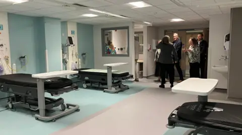 A room with several beds in it. The beds are on trolleys and there are a group of people by the room entrance 