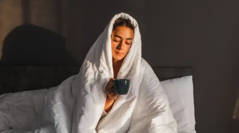 A woman holds a mug in bed, with her head covered by a blanket
