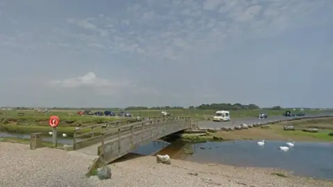 Google View from the shingle beach towards the road where there are about a dozen vehicles and and an ice-cream van parked. Between the beach and the road is a footbridge and a river where five swans are swimming.