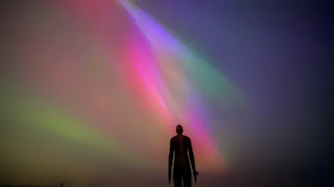 PA Media An iron statue with its arms by its sides is pictured in silhouette underneath the glorious pinks, greens and blues of the Northern Lights.