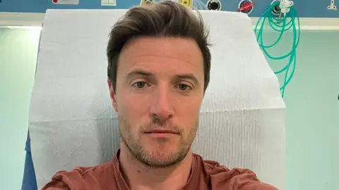 A man in a brown T-shirt takes a selfie in a hospital bed. He is not smiling.