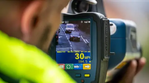 a Police officer wearing a high viz jacket holds a speed detection camera. On screen are a number of vehicles and data relating to their speed