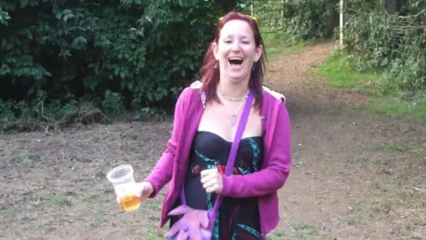 Amber Richards A woman with long brown hair and wearing a black dress and pink cardigan laughs while looking at the camera. There is woodland behind her, and she's holding a half-drunk pint glass