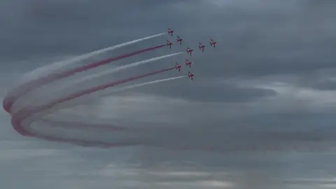 The Red Arrows against a cloudy sky with red and white smoke looping back behind them.