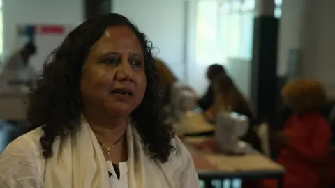Poornima Kirloskar-Saini wearing a white shirt and white shawl sitting in a classroom with women using sewing machines behind her