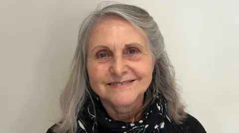 John Devine/BBC Sue Lowe is in the middle of the image smiling. She has long grey hair and is wearing a black T-shirt and a black and white scarf. She is stood in front of a grey wall. 