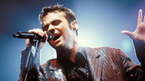 Getty Images Marti Pellow from Wet Wet Wet singing into a microphone. He has short, dark hair and is wearing a shiny black shirt and a brown sequined jacket