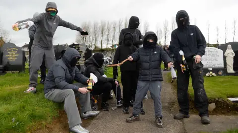 PA Media A group of people dressed in dark clothing are gathered in a graveyard. They all have their faces covered. Some are holding glass bottles, one is holding a golf club and another a stick.
