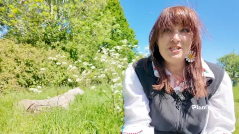 A woman with long hair and ear rings in the shape of daisies examines the flowers in the long grass around her
