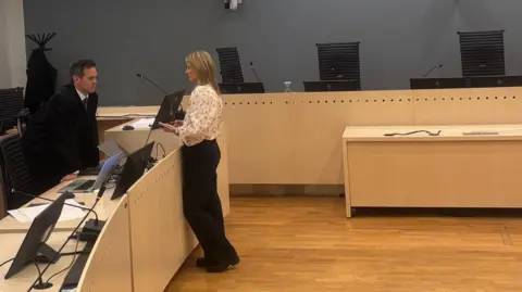 Paul Kirby/BBC A man to the left talks to a woman in a courtroom - he wears a suit, she wears a light coloured blouse