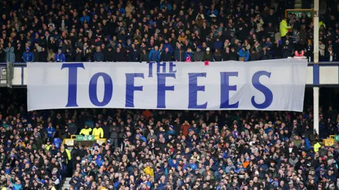 PA Media massive white banner with The Toffees branded on it in blue is draped across an upper stand with fans packed above and in the stand below at a match