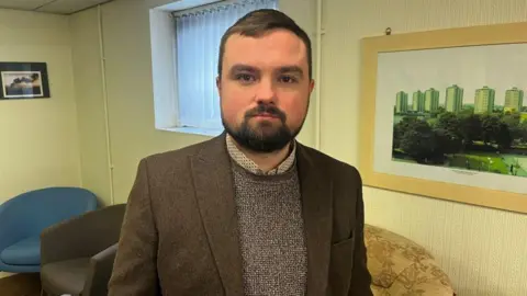 BBC /Anna Jameson A man with short dark hair, looks at the camera while standing in a room with chairs behind him