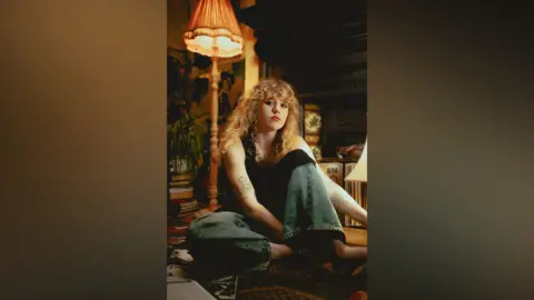 MGMT Press Shot Publicity shot of headline act Fiona-Lee. She is sitting on the floor in a dimly lit room surrounded by old fashioned furniture, including a tall lamp with a frilly shade. She has long wavy hair and is wearing a black top and jeans.