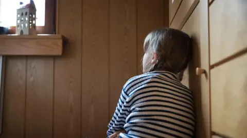 Getty Images A young girl sits with her back to the camera. She is leaning against a wooden door panel. She wears a black and white striped top and blue jeans. She has long brown hair and looks up to a window. There is a small model of a white house on the window sill. 