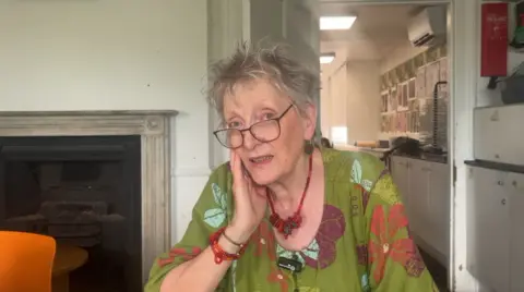 Jane Atkinson is sat in a chair resting her chin in her hand. She wears a green floral blouse, chunky red necklace and bracelets and glasses. There is a fireplace behind her.
