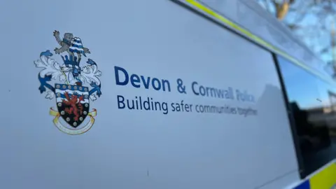 The view of the side of a police van. It has the words, in blue, 'Devon and Cornwall Police' on the side of the white van. 