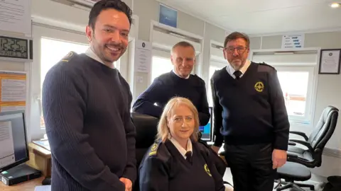 Four coastwatch volunteers in uniform standing together and looking at the camera. They are standing in the station cabin and smiling at the camera.