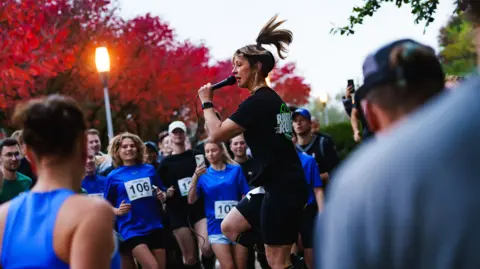 Ravers2Runners Jo, wearing black running gear and standing with a microphone in her hand looking at a group of runners with race numbers bibs on, getting them warmed up before a run