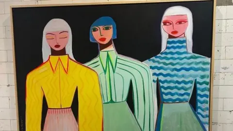 Margo in Margate A painting hanging on a tiled wall. Set against a black background, one is dressed in vibrant yellow, one is in green and another is in blue.