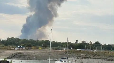 Huge plumes of smoke rising above a line of trees, which are by a riverbed with boats on it.