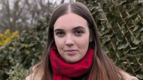 Molly looks straight ahead, standing in front of greenery – she has long brown hair parted in the middle and is wearing a red scarf