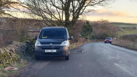 Two vehicles parked on the sides of a dry stone wall-lined rural road