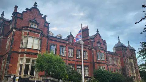 Anttoni James Numminen/LDRS Scarborough Town Hall is shown, a large building made of red bricks with a large number of windows across the front. The building also has multiple small spires. In the foreground, there are green trees, a black spiked fence and a tall flagpole displaying a Union Flag