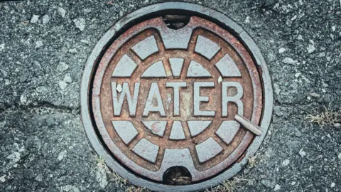 Getty Images A metal water main cover on a road.