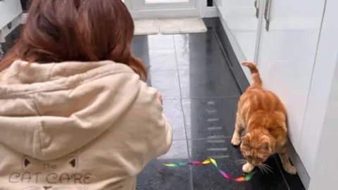 Sheree is crouched down on a dark grey tiled floor in a kitchen playing with a ginger cat