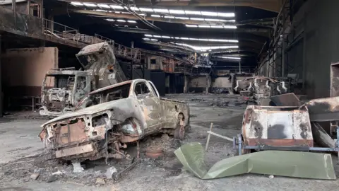 SAM READ/BBC This shows a warehouse building severely damaged by fire. There are holes in the roof allowing daylight through. In the foreground the remains of a burnt- out pick-up truck can be seen. Behind it a burnt-out lorry. Other burnt-out items can be seen to the right of the picture. The floor is covered in debris and in the background it looks like part of the roof has fallen in.