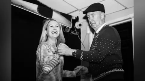 Fern Deacon/Stare Society Studio A woman with long blonde hair is smiling while dancing with an older man wearing a dark-coloured cap. It is a black and white photo.