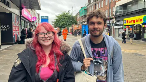 A young woman and young man looking at the camera with a shopping street behind them