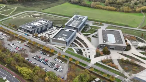 An aerial view of the Wellcome Genome Campus which is located next to a road and fields. There are four large buildings and several walkways. There is a car park in front of the campus to the left.