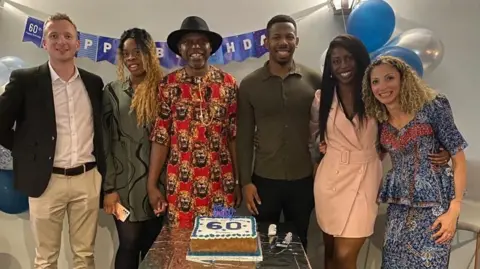 Supplied A family photo showing six people smiling as they stand behind a small table with a cake on it. The icing on the cake reads "60", while a blue and white banner reads "Happy Birthday". There are blue and silver balloons. An older man, wearing a black fedora hat and a long red shirt with lion-print design, stands in the centre holding hands with a younger woman, who is wearing a green dress, to the left. On the far left, a man wearing a black suit, white shirt and cram slacks, stands with his arms behind his back. To the right stand a younger man, wearing a green shirt, and another younger woman, wearing a pink dress. On the far right stands a woman with curly blond hair, who is wearing a blue patterned top and matching skirt.