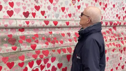 Alan Handley Alan Handley pictured looking at a stone wall with pink and red drawn hearts. He is bald and wearing dark tinted glasses and a dark blue coat.