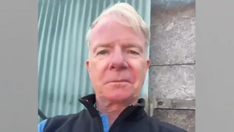Edward, a man with grey hair and blue eyes. He is wearing a navy shell jacket over a blue t shirt.