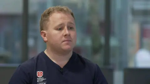 The picture shows trainee doctor David McCleary who has short fair hair and is wearing a navy v-neck medical shirt
