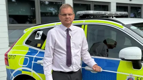 Essex Police Adam Pipe wears a white shirt and purple patterned tie. He leans against the door of a yellow and blue marked police car.