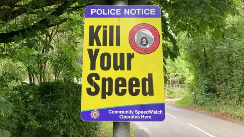 A blue and yellow road sign reading "kill your speed", "Community SpeedWatch Operates Here" pictured on a tree-lined, country road.
