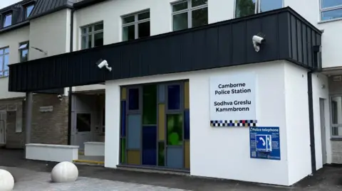 The black and white front of the newly refurbished public enquiry office at Camborne police Station, showing information signs on the outside and a large coloured glass window in the middle of the building.